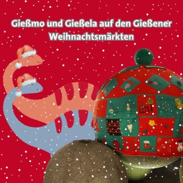 Zwei kindliche Dinosaurier, einer rot, einer blau, stehen hinter einer Kugelskulptur. Sowohl die Dinosaurier, als auch eine der Kugeln haben Weihnachtsmützen auf.