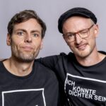 Kopf und Oberkörper zweier Personen, die in die Kamera schauen. Sie haben je einen Arm um den anderen gelegt. Beide schauen in die Kamera- Eoner hat Mütze und Brille auf und zwinkert.