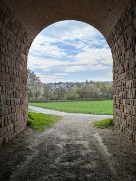 Blick durch einen Tunnel auf ein grünes Feld, im Hintergrund Bäume und dahinter eine Ortschaft. Der Himmel ist blau mit weißen Wolken.