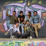 Eine Gruppe von 6 Personen posieren vor einer mit Grafitti besprühten Wand