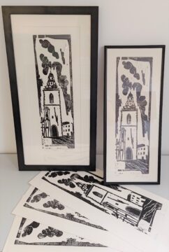 Mehrere Drucke des gleichen Kirchenmotivs sind zu sehen. Zwei davon sind eingerahmt, einer im Hochformat mit dickem schwarzen Rahmen, ein anderer etwas schmaler mit dünnem Rahmen. Davor liegen mehrere weitere ungerahmte Drucke auf dem Tisch.