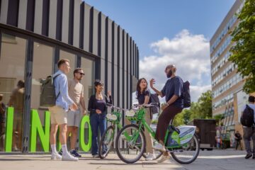 Eine Person auf einem Fahrrad, vier weitere stehend vor einem modernen Gebäude